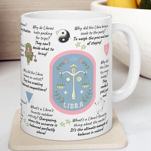 May include: White ceramic mug with a colorful design featuring the Libra zodiac sign, scales, and text about Libra personality traits. The text includes phrases like "Why do Libras hate packing for trips?" and "What's a Libra's favorite outdoor activity? Stargazing."
