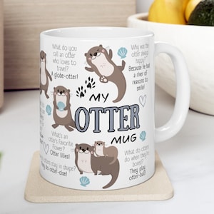 Pode incluir: Caneca de cerâmica branca com um design envolvente com lontras de desenho animado, trocadilhos e o texto "My Otter Mug". As lontras são retratadas em várias poses com conchas e pegadas. A caneca está em uma base clara.