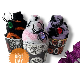 Puede incluir: Tres regalos con forma de cupcake con temática de Halloween. Cada uno tiene una araña decorativa y una gema. Las magdalenas son negras y naranjas, rosas y blancas, y moradas y negras. Un murciélago morado brillante está al lado de las magdalenas.