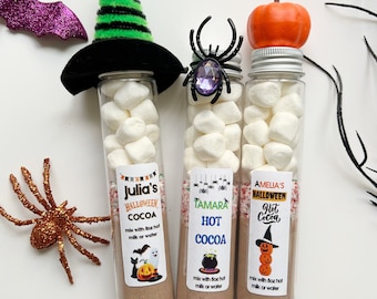 May include: Three glass tubes filled with hot cocoa mix and marshmallows, decorated with Halloween-themed toppers. The tubes are labeled "Julia's Halloween Cocoa", "Tamara Hot Cocoa", and "Amelia's Halloween Hot Cocoa".