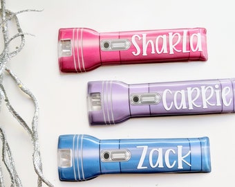Puede incluir: Tres linternas de colores, cada una con un nombre diferente: Shakla, Carrie y Zack. Las linternas son de color rosa, morado y azul. Cada linterna tiene un botón plateado y una lente transparente.