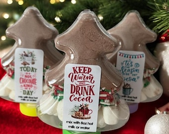 Può includere: Tre contenitori a forma di albero pieni di miscela di cacao e marshmallow. Ogni contenitore ha un'etichetta con testo come "Keep Warm and Drink Cocoa". I contenitori sono su una superficie rossa.