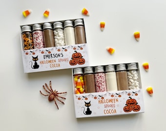 May include: Two boxes of Halloween Spooky Cocoa with glass vials filled with ingredients. Each box has a black cat, pumpkins, and the text "Emerson's Halloween Spooky Cocoa". Candy corn and a glitter spider are scattered around.