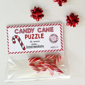 Puede incluir: Una bolsa transparente con bastones de caramelo rojos y blancos, con una etiqueta que dice "CANDY CANE PUZZLE" y el nivel "Intermedio". Tres lazos decorativos rojos est&aacute;n colocados alrededor de la bolsa. El rompecabezas es para mayores de 3 a&ntilde;os.