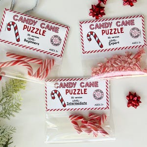 Puede incluir: Tres bolsas transparentes con rompecabezas de bastones de caramelo, cada una etiquetada como "Candy Cane Puzzle" con niveles de dificultad: Principiante, Experto e Intermedio. Los rompecabezas son para mayores de 3 a&ntilde;os.