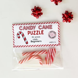 Puede incluir: Una bolsa transparente con bastones de caramelo a rayas rojas y blancas y un paquete de "Candy Cane Puzzle". El paquete tiene un borde rojo y blanco y presenta una ilustraci&oacute;n de bast&oacute;n de caramelo y el texto "3D version, Level: Beginners, Ages 3+". Tres lazos rojos est&aacute;n dispersos.