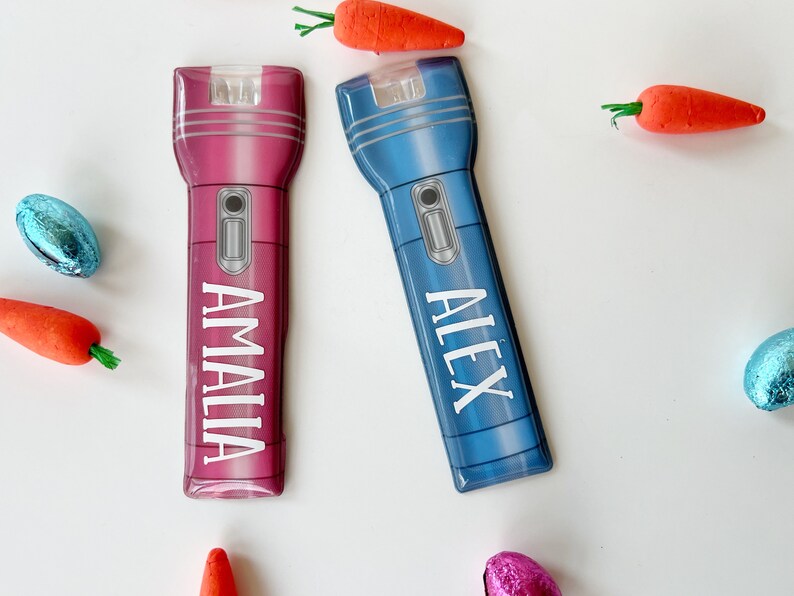 Peut inclure: Deux lampes de poche personnalis&eacute;es, une rose et une bleue, chacune avec un nom imprim&eacute; verticalement. La lampe de poche rose dit "AMALIA" et la bleue dit "ALEX". Les lampes de poche sont entour&eacute;es de d&eacute;corations de carottes oranges et d'&oelig;ufs en chocolat emball&eacute;s.