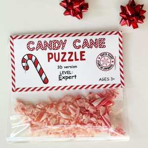 Puede incluir: Una bolsa de pl&aacute;stico transparente llena de bastones de caramelo rojos y blancos triturados, con una etiqueta que dice "CANDY CANE PUZZLE". La etiqueta tambi&eacute;n incluye un gr&aacute;fico de bast&oacute;n de caramelo, el texto "3D version", "LEVEL: Expert" y "AGES 3+". Tres lazos decorativos rojos est&aacute;n dispersos alrededor del rompecabezas.