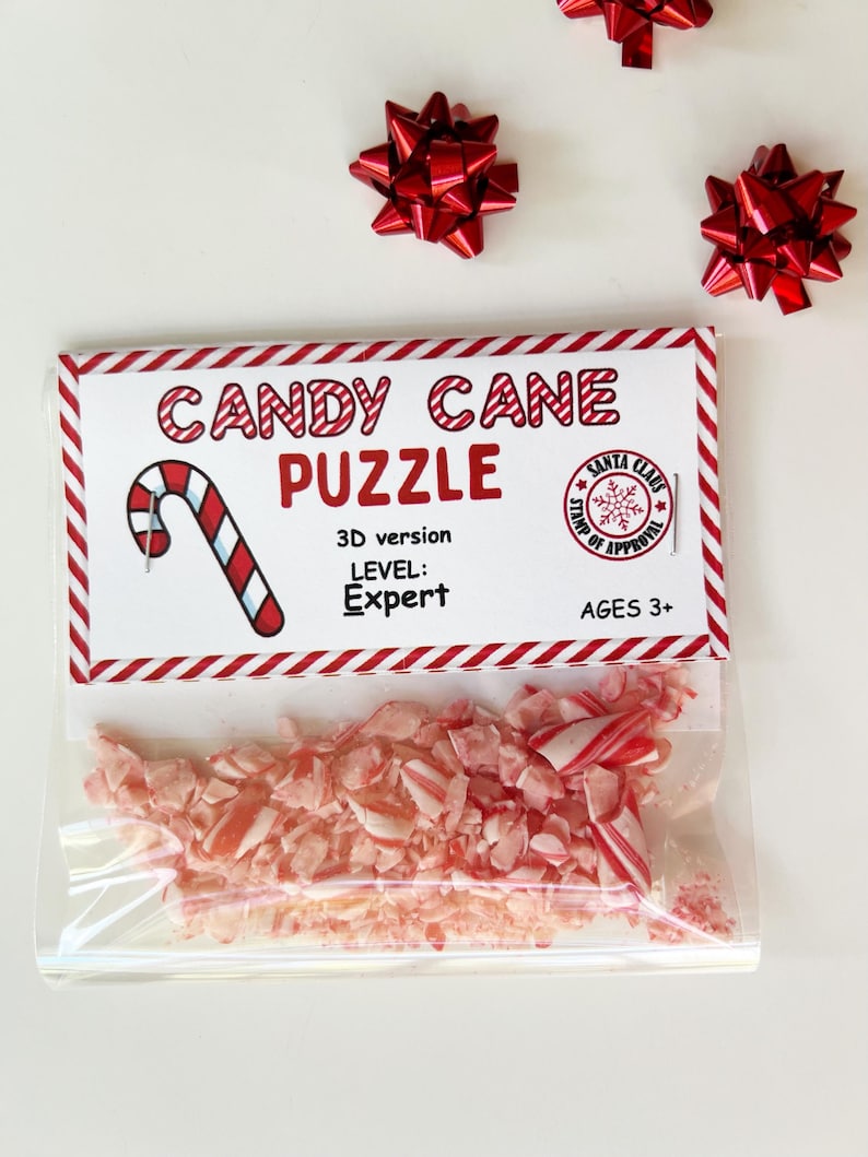 Puede incluir: Una bolsa de pl&aacute;stico transparente llena de bastones de caramelo rojos y blancos triturados, con una etiqueta que dice "CANDY CANE PUZZLE". La etiqueta tambi&eacute;n incluye un gr&aacute;fico de bast&oacute;n de caramelo, el texto "3D version", "LEVEL: Expert" y "AGES 3+". Tres lazos decorativos rojos est&aacute;n dispersos alrededor del rompecabezas.