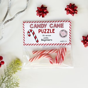 Puede incluir: Una bolsa de pl&aacute;stico transparente que contiene tres bastones de caramelo a rayas rojas y blancas, con una etiqueta que dice "Candy Cane Puzzle" y el nivel "Principiante". La imagen est&aacute; decorada con lazos rojos y vegetaci&oacute;n festiva.