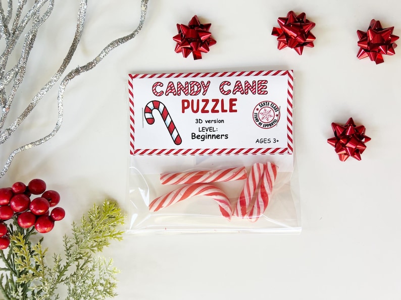 Puede incluir: Una bolsa de pl&aacute;stico transparente que contiene tres bastones de caramelo a rayas rojas y blancas, con una etiqueta que dice "Candy Cane Puzzle" y el nivel "Principiante". La imagen est&aacute; decorada con lazos rojos y vegetaci&oacute;n festiva.