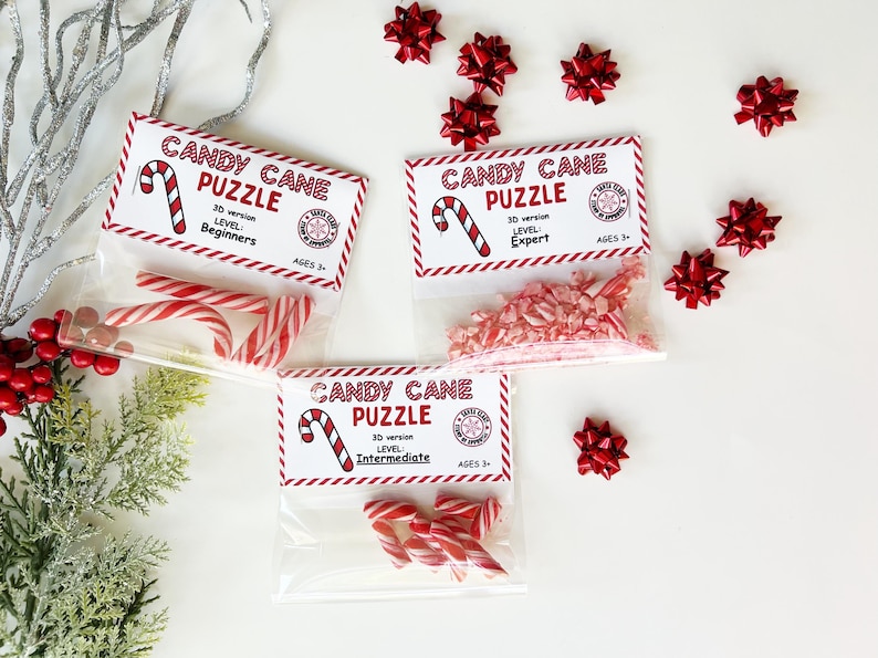 Puede incluir: Tres bolsas transparentes con rompecabezas de bastones de caramelo, cada una con una etiqueta que dice "Candy Cane Puzzle" y niveles de dificultad: Principiante, Experto e Intermedio. Las bolsas contienen bastones de caramelo a rayas rojas y blancas y caramelos triturados. Hay lazos rojos esparcidos.