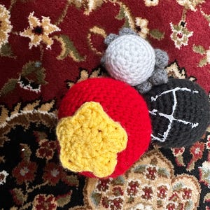 May include: Three crocheted spheres: a large red ball with a yellow five-pointed star attached to the front, a smaller light gray ball with textured surface details, and a black ball featuring a white embroidered letter H on its side.