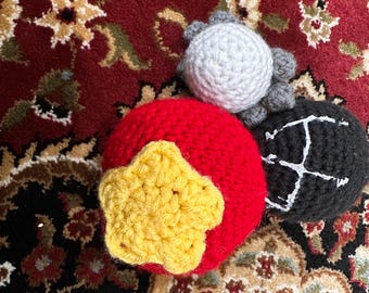 May include: Three crocheted spheres: a large red ball with a yellow five-pointed star attached to the front, a smaller light grey ball with textured surface details, and a black ball featuring a white embroidered letter H on its side.