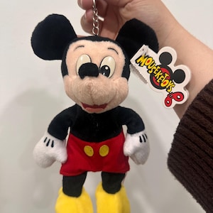 May include: A Mickey Mouse plush keychain. The toy features black ears, a black body, red shorts with yellow buttons, yellow shoes, and white gloves. The keychain has a silver metal clasp and a tag that reads "MOUSEKETOTS".