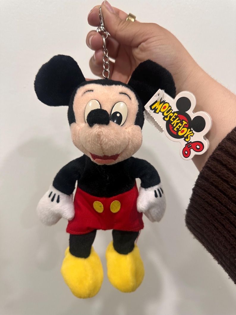 May include: A Mickey Mouse plush keychain. The toy features black ears, a black body, red shorts with yellow buttons, yellow shoes, and white gloves. The keychain has a silver metal clasp and a tag that reads "MOUSEKETOTS".