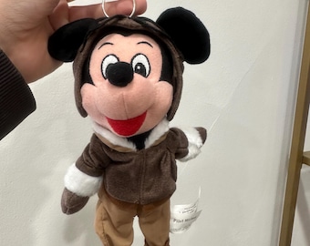 May include: A plush Mickey Mouse keyring dressed as a pilot, featuring a brown aviator cap, a brown jacket with white faux fur trim, and tan trousers. The character has black ears, a pink face, and a red mouth. A metal ring and clip are attached.