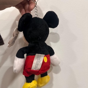 May include: A Mickey Mouse plush keychain. The toy features a black head and ears, white gloves, red shorts, and yellow shoes. The keychain is attached to the top of the head with a silver chain.