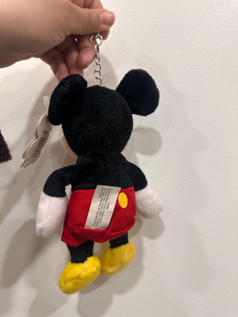 May include: A Mickey Mouse plush keychain. The toy features a black head and ears, white gloves, red shorts, and yellow shoes. The keychain is attached to the top of the head with a silver chain.