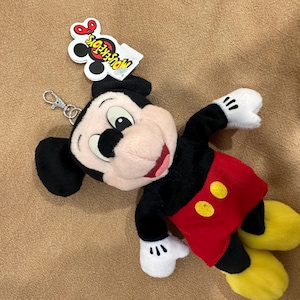 May include: A Mickey Mouse plush keychain. The toy has a black body, red shorts with yellow buttons, and yellow shoes. The keychain has a silver clasp and a tag that reads "MOUSE TOYS".