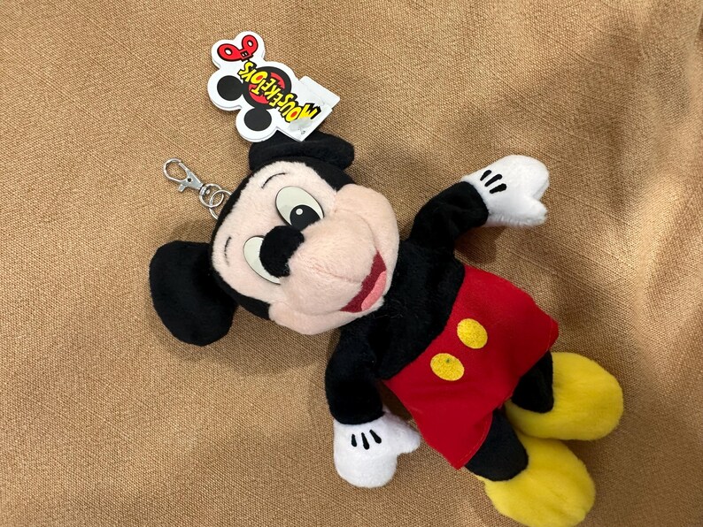 May include: A Mickey Mouse plush keychain. The toy has a black body, red shorts with yellow buttons, and yellow shoes. The keychain has a silver clasp and a tag that reads "MOUSE TOYS".