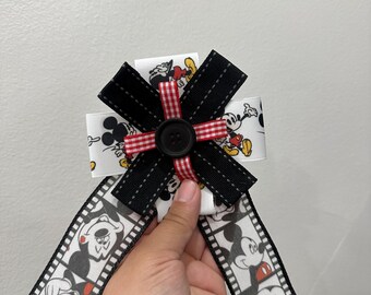 May include: A decorative hair bow featuring white ribbon with a repeating Mickey Mouse print, layered with black ribbon strips and a red and white gingham ribbon cross, centred with a black button and two long ribbon tails with film strip graphics.