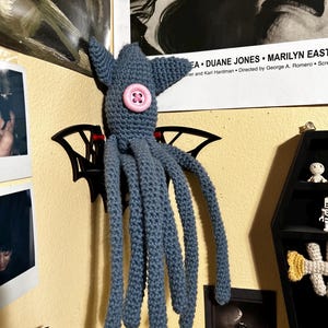 Può includere: Un polpo blu all'uncinetto fatto a mano con un bottone rosa come occhio. Il polpo è montato su una decorazione murale nera a forma di pipistrello. L'immagine è ambientata contro un muro giallo con foto incorniciate e un poster di film.