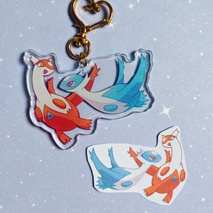 Peut inclure: Porte-clés en acrylique transparent avec deux personnages de dessins animés, l'un orange et blanc, l'autre bleu et blanc, avec un fermoir en forme de cœur doré. Un autocollant plus petit du même motif est en dessous. Le fond est bleu clair avec des étoiles blanches.