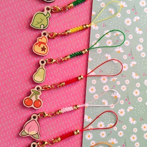 Puede incluir: Una colección de colgantes de teléfono coloridos con varios diseños de frutas e iconos. Cada colgante tiene un cierre dorado y un cordón trenzado en colores como azul, verde, amarillo, rojo y naranja. El fondo es un patrón floral rosa y verde.