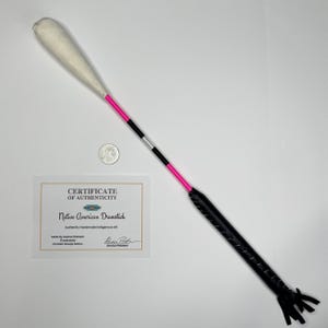 Könnte beinhalten: Ein indianischer Trommelstock mit einem weiß gepolsterten Kopf, einem rosa, schwarzen und silbernen Schaft und einem schwarzen Ledergriff mit Fransen. Ein Echtheitszertifikat und eine Münze sind ebenfalls im Bild.