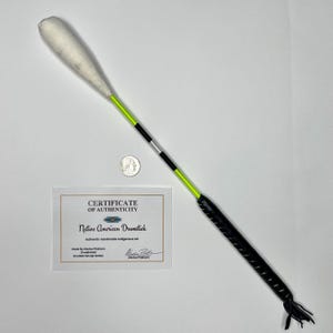 Könnte beinhalten: Ein indianischer Trommelstock mit einem weiß gepolsterten Kopf, einem neongrünen und schwarzen Schaft und einem schwarzen Ledergriff. Das Echtheitszertifikat lautet "Native American Drumstick".