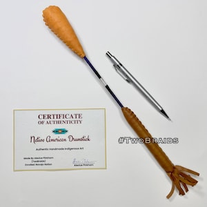 Könnte beinhalten: Zwei indianische Trommelschlägel mit hellbraunen Lederköpfen und -griffen, einer mit Fransen. Ein Echtheitszertifikat ist vorhanden, zusammen mit einem silbernen Druckbleistift. Das Zertifikat lautet "Native American Drumstick".