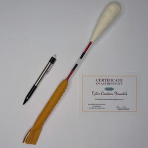 May include: A drumstick with a textured cream-colored head, a red shaft with black and silver bands, and a tan leather-wrapped handle with fringe. Beside it lies a silver pen and a Certificate of Authenticity for a Native American Drumstick.
