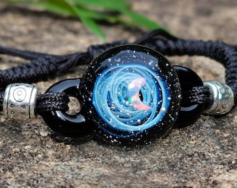May include: A black cord bracelet with a round glass bead featuring a swirling blue and white galaxy design with a pink crescent moon.
