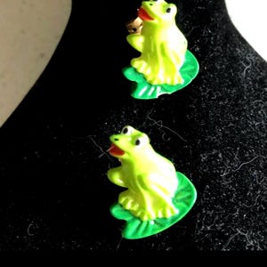 May include: A pair of green frog earrings with red mouths and gold accents. The frogs are sitting on green lily pads.