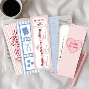 May include: A collection of decorative bookmarks with various designs, including stripes, floral patterns, and text. One bookmark reads "Pretty Girls Books," another "The Romance Readers Edition," and a third says "Book Lover." A cup of coffee and a pen are also present.