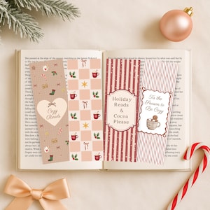 Puede incluir: Un libro abierto con varios marcapáginas festivos. Los marcapáginas presentan diseños navideños con el texto "Cozy Reads", "Holiday Reads & Cocoa Please" y "Tis the Season to Be Cozy". También se ve un bastón de caramelo y un lazo.