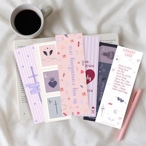 Könnte beinhalten: Eine Sammlung dekorativer Lesezeichen mit verschiedenen Designs und Texten, darunter Sätze wie "in my romantasy era" und "Book Friends". Die Lesezeichen zeigen Illustrationen von Herzen, Kronen, Schwertern und Flügeln, mit einem rosa Stift und einer Tasse Kaffee.