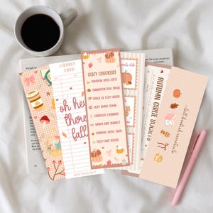 Könnte beinhalten: Eine Sammlung von Lesezeichen zum Thema Herbst mit verschiedenen Designs, darunter Kürbisse, Blätter und dem Text "Oh hey there fall". Ein Lesezeichen listet eine "Cozy Checklist" auf. Eine Tasse Kaffee und ein rosa Stift sind ebenfalls zu sehen.