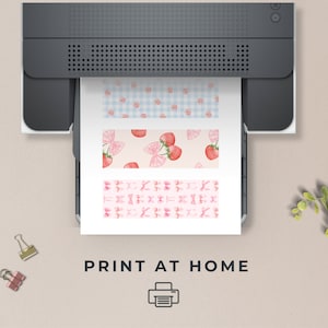 May include: A dark gray printer outputting a white sheet of paper featuring three horizontal strips with patterns. The top strip has blue gingham with strawberries, the middle has strawberries and pink bows, and the bottom has pink bows. Text reads PRINT AT HOME.