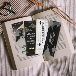 May include: Four black and white bookmarks with different designs. The bookmarks feature dragons, clouds, and text. The text on the bookmarks reads "Dragon Riders Society", "Shadow Daddy", "A Reader Without Their Book Is A Tragedy", and "Ride or Die".