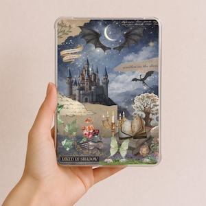 Pode incluir: Uma capa transparente para tablet com um design de fantasia. A arte inclui um castelo, asas de dragão, uma lua crescente e os textos "enemies to lovers" e "written in the stars". A capa tem uma estética vintage com detalhes florais e borboletas.