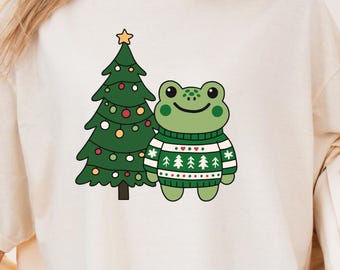 May include: Cream-colored t-shirt featuring a cartoon frog wearing a green and white sweater, standing next to a decorated Christmas tree. The tree has a gold star on top and colorful ornaments. A festive design for the holiday season.