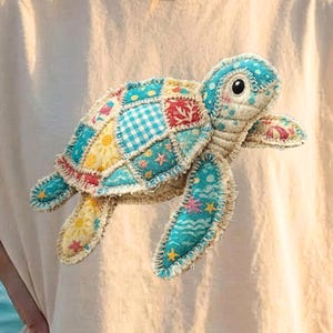 Puede incluir: Una camiseta beige con un diseño de tortuga de retazos coloridos. La tortuga tiene una cabeza azul, un caparazón estampado y aletas azul claro. La camiseta está hecha de un material suave y es adecuada para uso casual. El fondo muestra un océano borroso.
