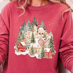 May include: A red-toned sweatshirt featuring a watercolor illustration of a snow-covered village with red, white, and yellow houses, evergreen trees, and a church. The design is centered on the chest.