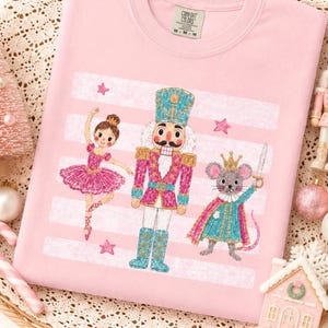 May include: A light pink crewneck t-shirt featuring a graphic print of a ballerina, a nutcracker, and a mouse king. The design includes horizontal white stripes and small pink stars. The shirt has a Comfort Colors brand label at the inner neckline.