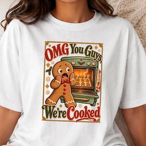 May include: A white short-sleeve t-shirt featuring a graphic of a shocked gingerbread person standing in front of an open oven with smaller gingerbread cookies inside. The text reads, "OMG You Guys, We're Cooked" in red and green lettering.