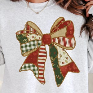 Puede incluir: Una sudadera gris claro con un gran diseño de lazo colorido. El lazo está compuesto por varias telas estampadas en rojo, verde y beige, con un contorno dorado. El diseño tiene una estética de patchwork, ideal para la temporada navideña.