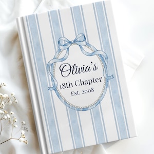 Pode incluir: Um livro de capa dura com um design de listras azuis e brancas. A capa apresenta uma fita azul e uma moldura oval com o texto "Olivia's 18th Chapter Est. 2008". O livro está sobre uma superfície branca com detalhes florais.