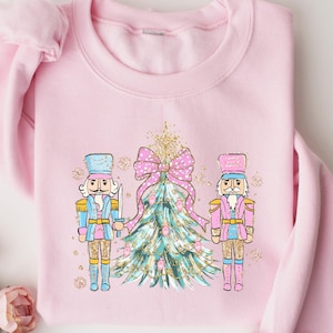 May include: Pink sweatshirt with a Christmas tree and two nutcracker soldiers. The tree is decorated with a pink bow and gold glitter. The nutcrackers are wearing blue and pink uniforms.
